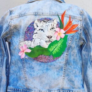 veste customisée panthère veste_en_jeans-customisee_pour_enfant_avec_dessin_de_panthere_et_de_fleurs_exotiques_vertes_et_oranges_sur_un_fond_rond_paillete_violet